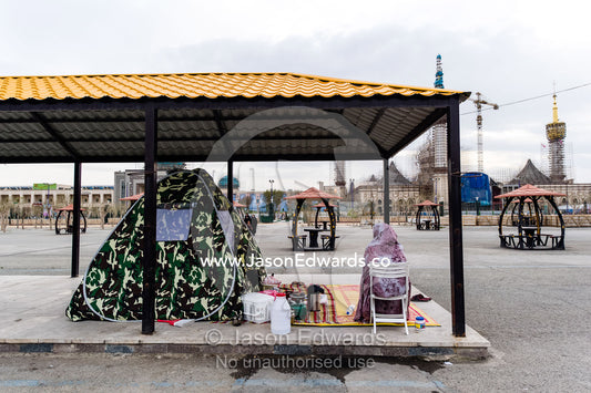 A woman camped beneath a bus shelter beside a mausoleum. Mausoleum of Ayatollah Khomeini, Tehran, Tehran Province, Islamic Republic of Iran.