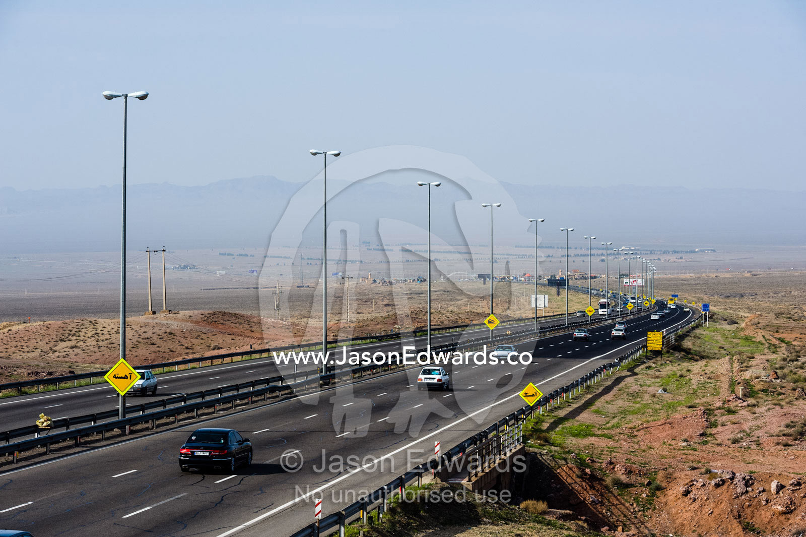 Traffic moves along a multi-lane highway crosses a flat plain in the desert. Qom Province, Islamic Republic of Iran.