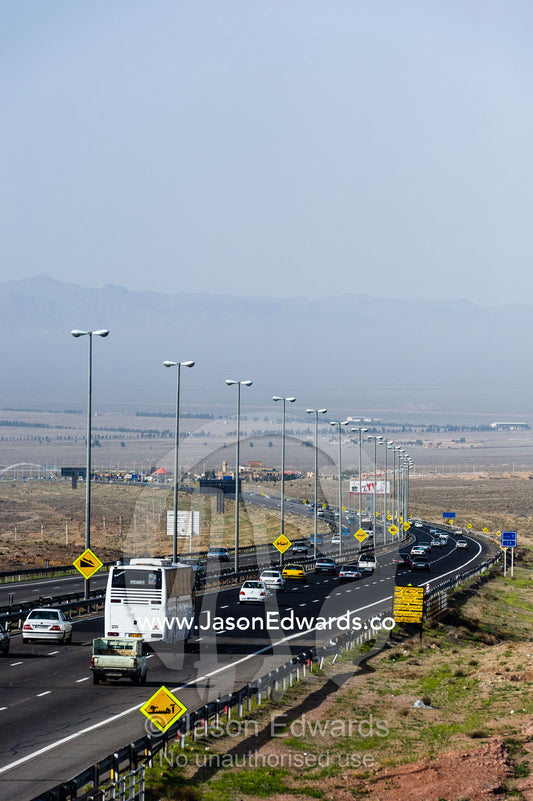 Traffic moves along a multi-lane highway crosses a flat plain in the desert. Qom Province, Islamic Republic of Iran.