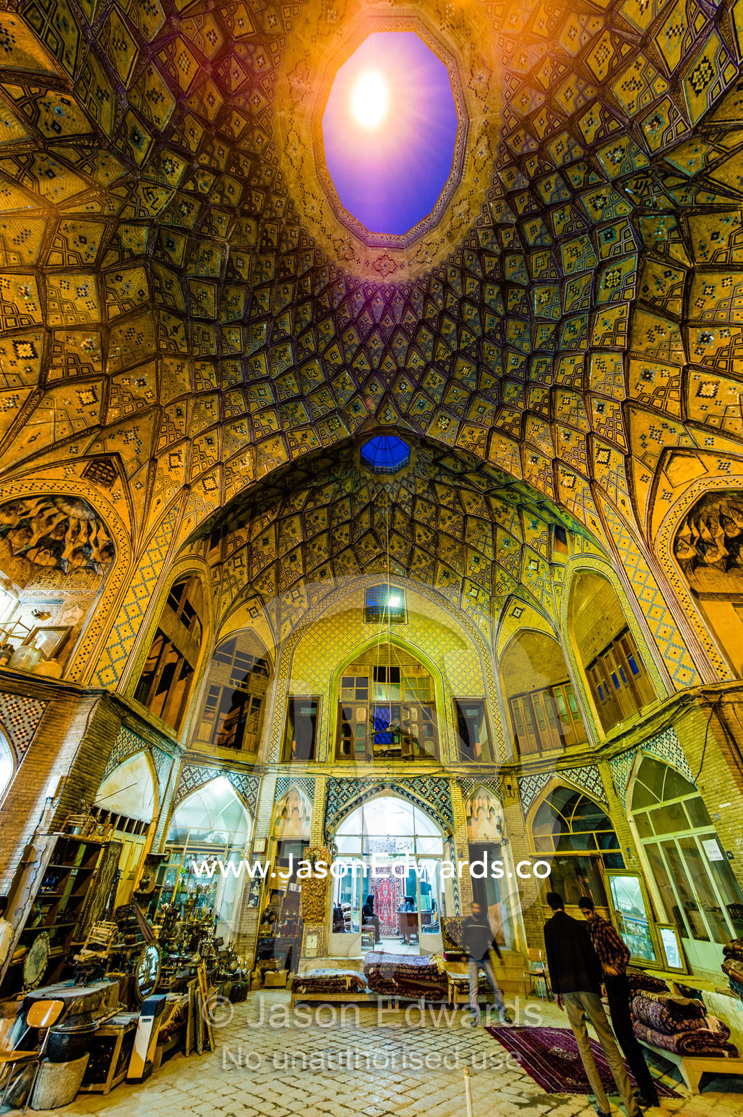 Carpet sellers and merchants display their wares beneath the vaulted spiralling geometric ceiling of an ancient bazaar. The Bazaar of Kashan, Kashan, Isfahan, Islamic Republic of Iran.