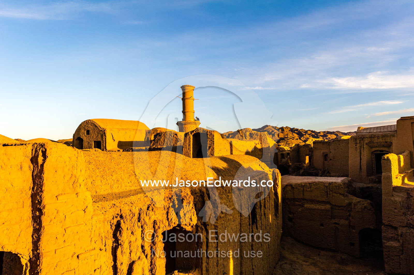 Sunset warms buildings of a mosque minaret and crumbling handmade brick ruins of an ancient abandoned village in the desert. Kharanagh, Yazd, Yazd Province, Islamic Republic of Iran.
