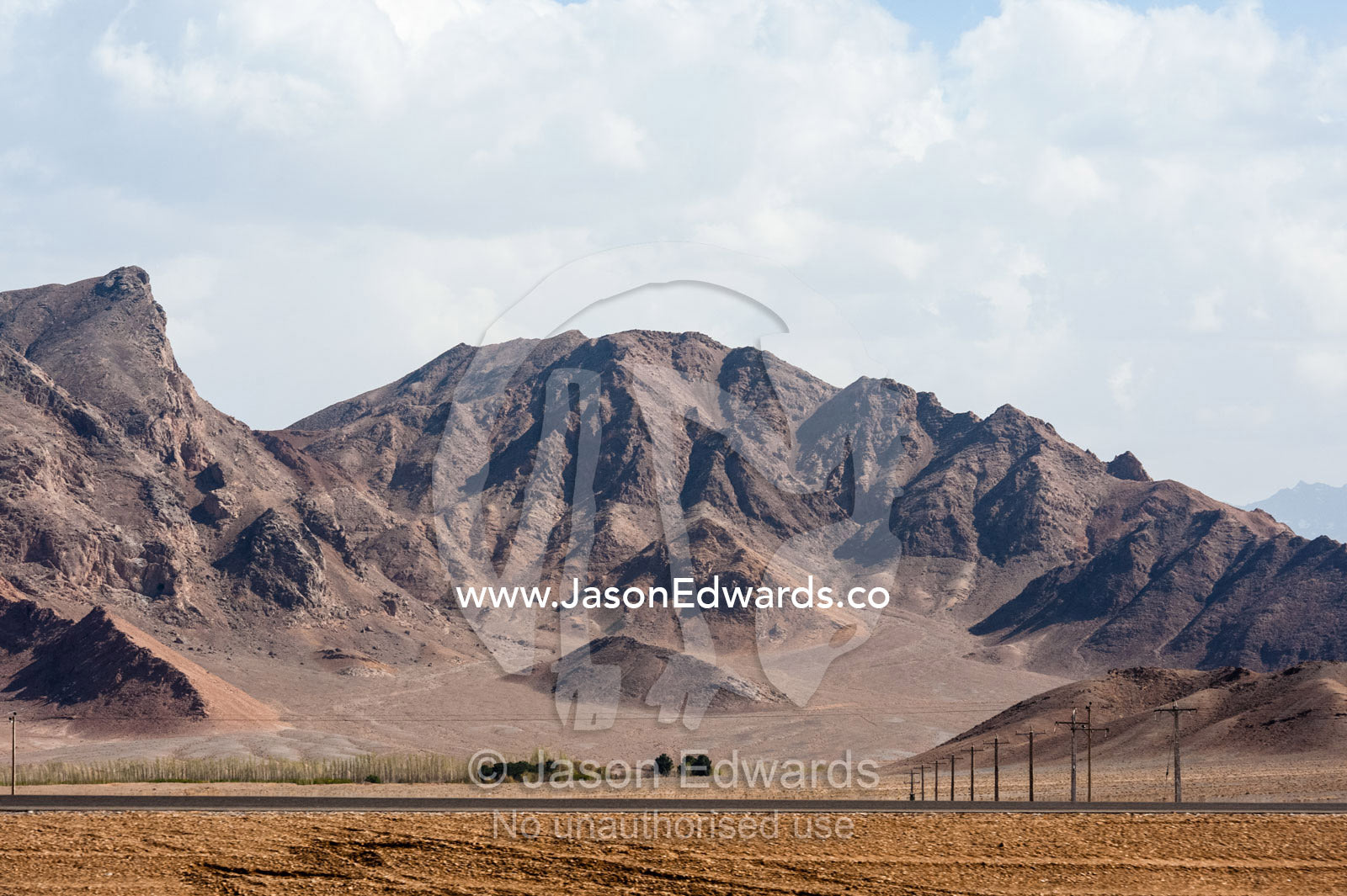 Telegraph poles and a flat desert highway beneath jagged mountain peaks. Shir Kuh Mountain, Yazd, Yazd Province, Islamic Republic of Iran.
