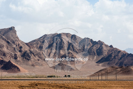 Telegraph poles and a flat desert highway beneath jagged mountain peaks. Shir Kuh Mountain, Yazd, Yazd Province, Islamic Republic of Iran.