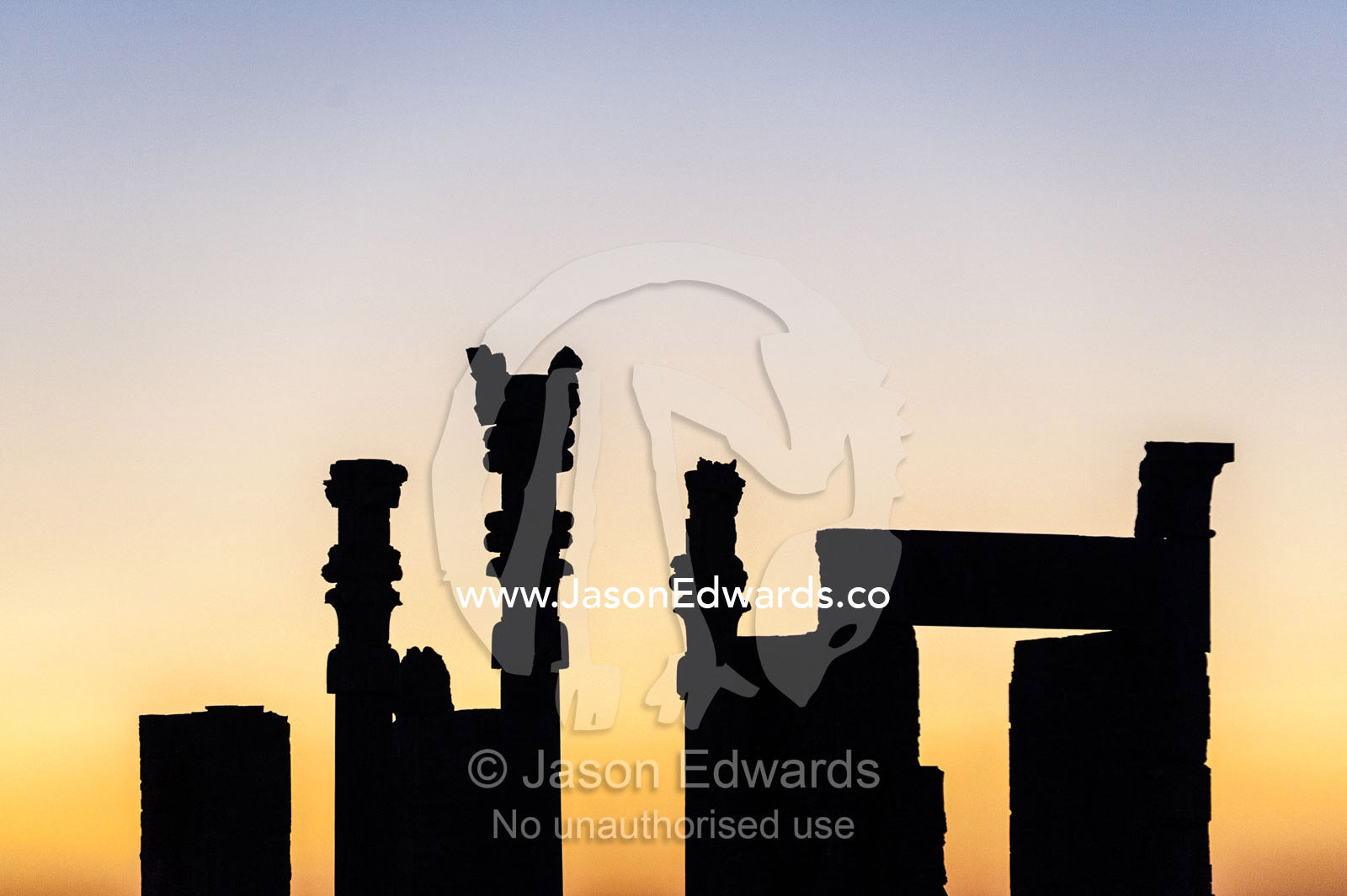 Stone columns and the Gate of All Nations silhouette against a darkening sky at sunset. Persepolis, Shiraz, Fars Province, Islamic Republic of Iran.