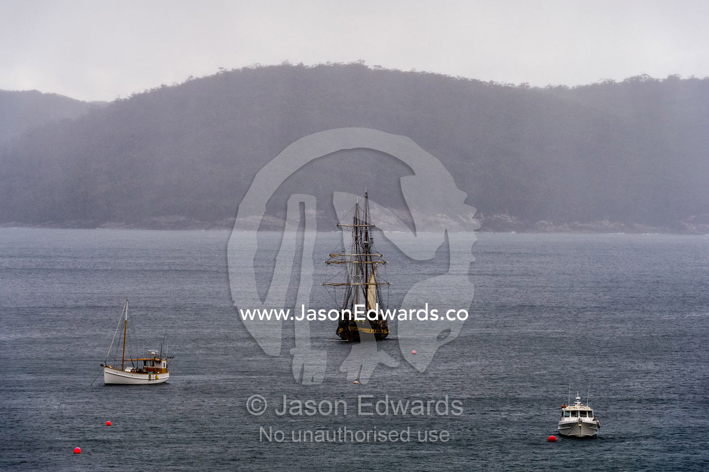 An antique sailing ship the Lady Nelson mooring in a bay on a rainy day. Mason Cove, Port Arthur Historic Site, Port Arthur, Tasmania,
Australia.