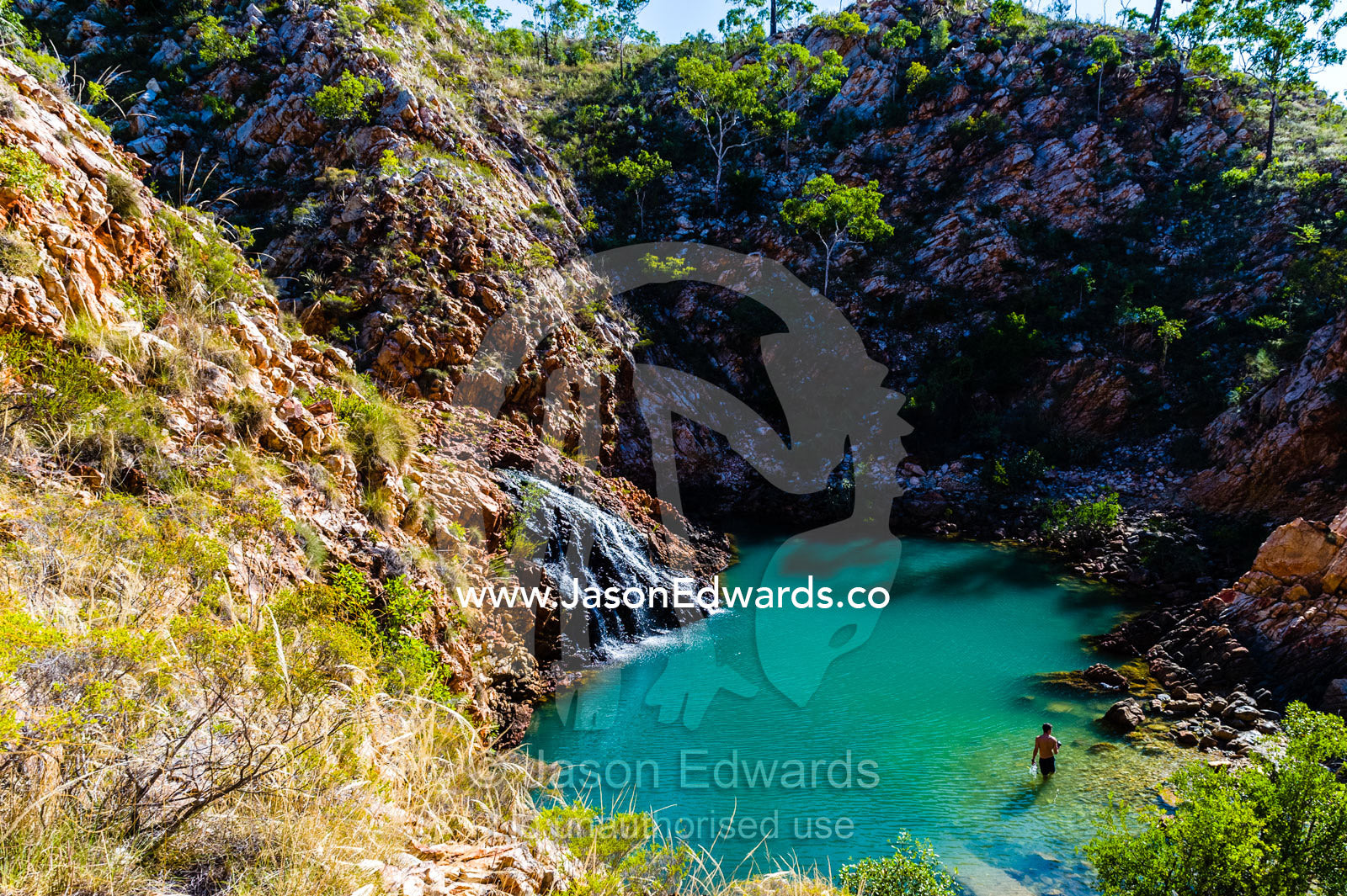 A man swimming in a small waterfall cascading into a sandstone gorge waterhole in the desert. Crocodile Creek, Kimberley Coast, Western Australia, Australia.