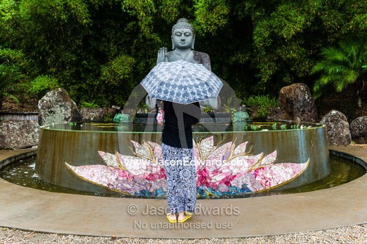 A tourist visits a statue of buddha offering a blessing over a lotus pond in a botanical garden. Crystal Castle, Shambhala Gardens, Mullumbimby, New South Wales, Australia.