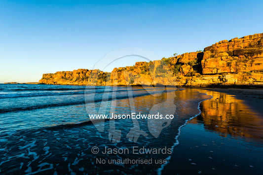 The reflection of orange ochre cliffs in waves lapping on a beach at sunset. Calamity Bay, Koolama Bay, Kimberley Coast, Western Australia, Australia.
