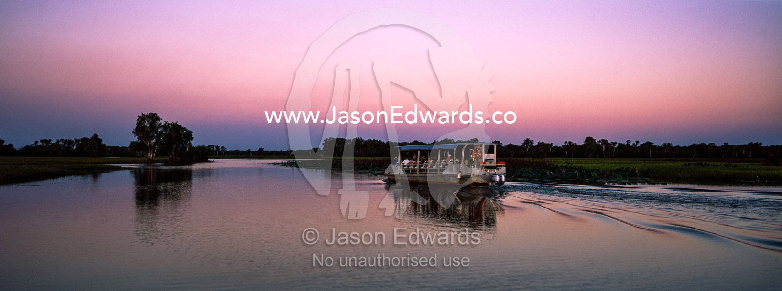 A tourist boat explores a tranquil billabong during the sunset afterglow. Yellow Water Billabong, Cooinda, Kakadu National Park, Northern Territory, Australia.