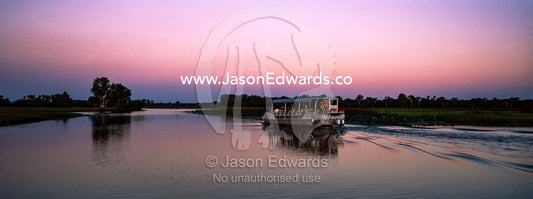 A tourist boat explores a tranquil billabong during the sunset afterglow. Yellow Water Billabong, Cooinda, Kakadu National Park, Northern Territory, Australia.