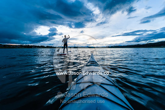 A canoe bow on a still lake and a man on a paddleboard beneath storm clouds. Peru.