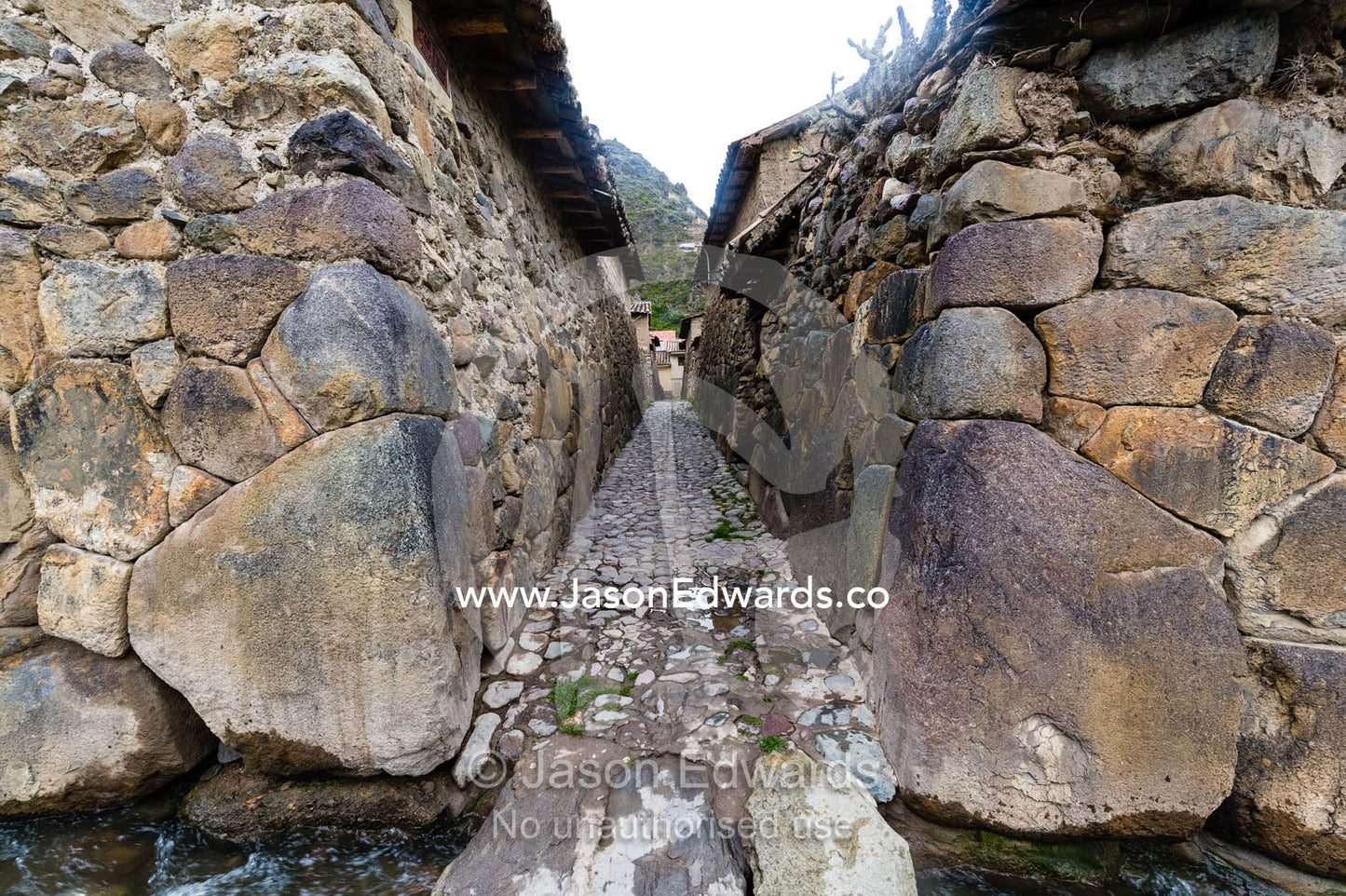 An irrigation canal in a village lane paved with stones and lined with high Inca dry-stone walls. Ollantaytambo, Peru.