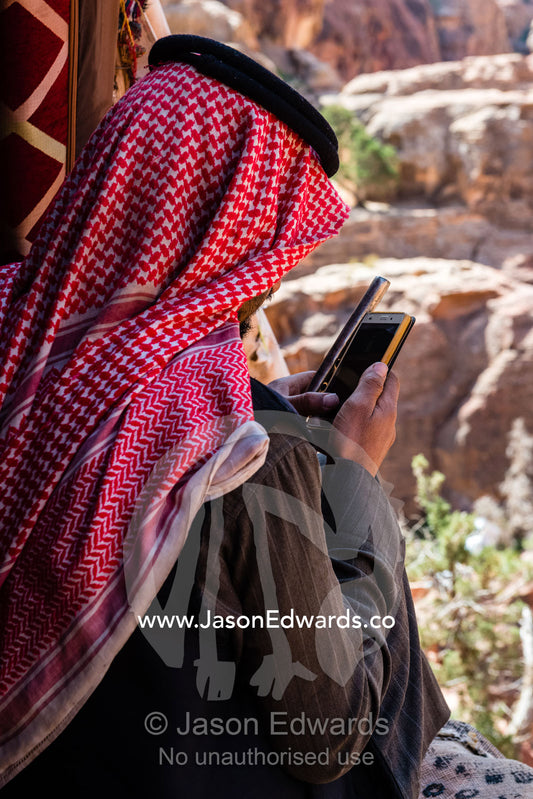 A Bedouin man wearing a keffiyeh, also known as ashemagh mhadab, using a smart phone above the Treasury in Petra. Petra, The Lost City of Petra, Jordan