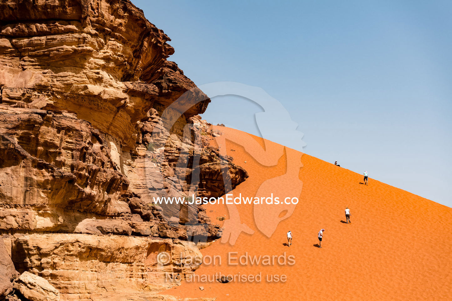 Tourists climb a sand dune washed up against a sandstone escarpment. Wadi Rum, The Valley of the Moon, Jordan