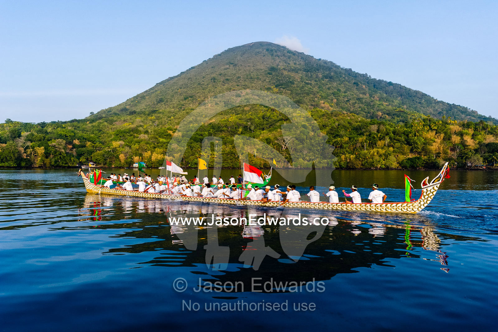 Villages paddling a long, decorated war canoe past an active volcano. Banda Neira, Gunung Api Volcano, Banda Island, West Papua, Indonesia