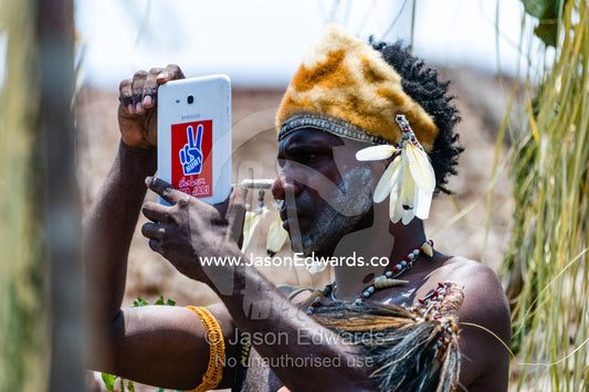 An Asmat warrior wearing a hat made of feathers, shells and Spotted Cuscus fur using a tablet device to record video. Syuru Village, Asmat Regency, Papua, Indonesia