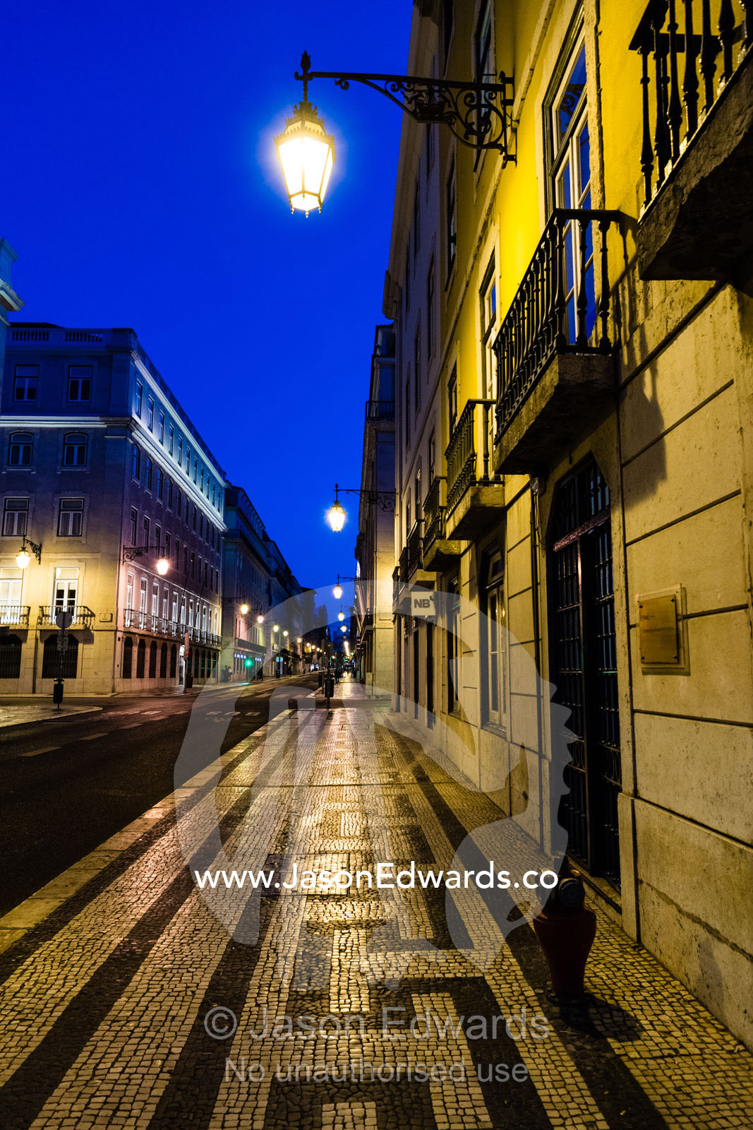 A tiled mosaics made from black basalt and white limestone pavers lines a sidewalk at night. Lisbon, Lisboa, Portugal