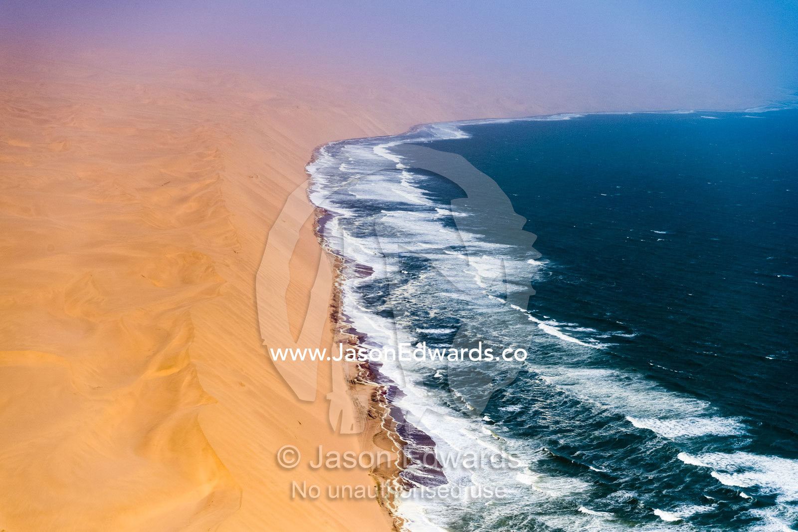 An aerial view of Atlantic Ocean waves pummeling enormous sand dunes along the desert coastline of Namibia. Namib Desert, Diamond Coast, Skeleton Coast, Namib-Naukluft National Park, Namibia