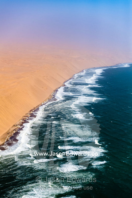 An aerial view of Atlantic Ocean waves pummeling enormous sand dunes along the desert coastline of Namibia. Namib Desert, Diamond Coast, Skeleton Coast, Namib-Naukluft National Park, Namibia