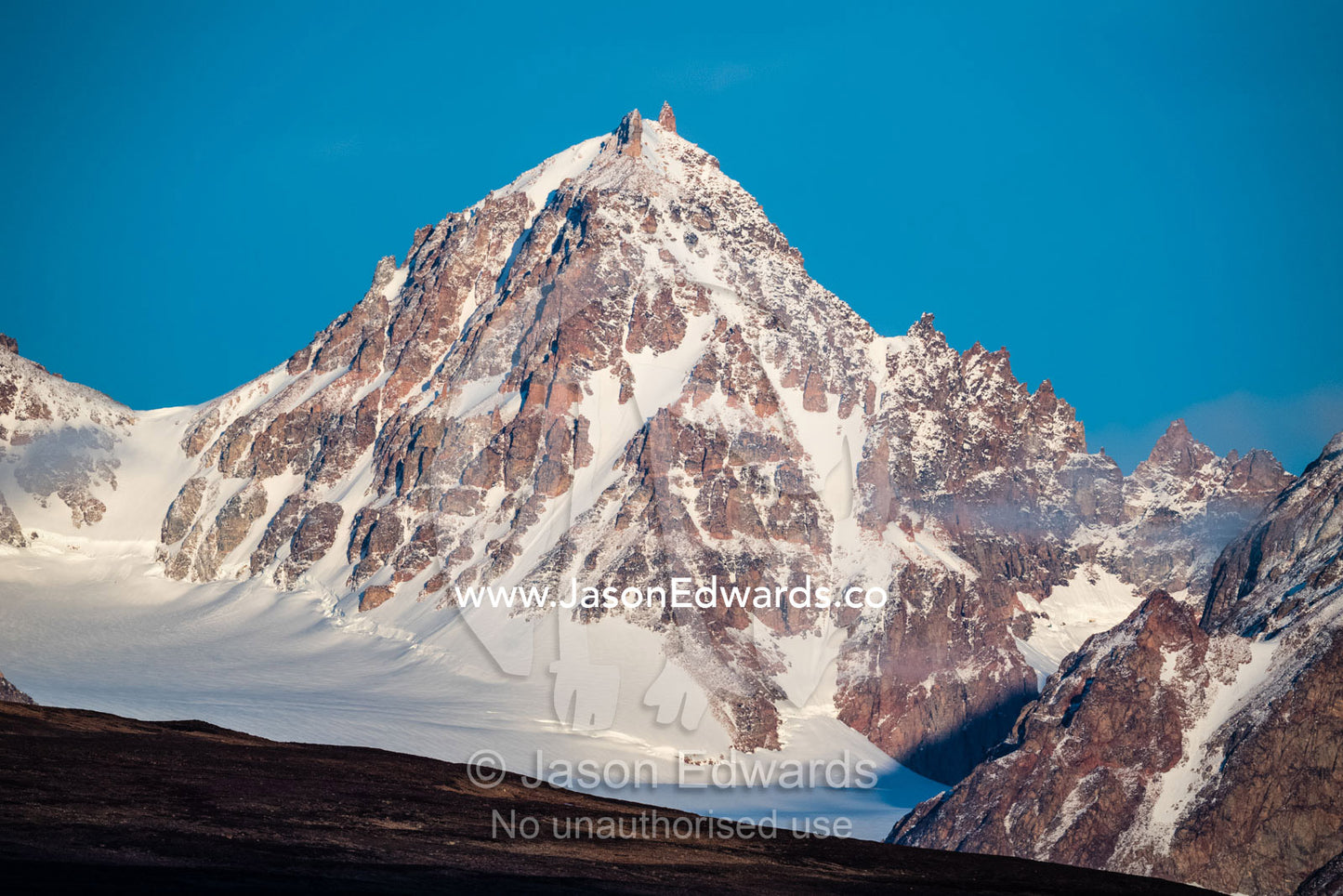 A jagged snow-covered mountain peak rises into a clear blue arctic sky. Constable Point, Hurry Inlet, Scoresby Sound, Greenland
