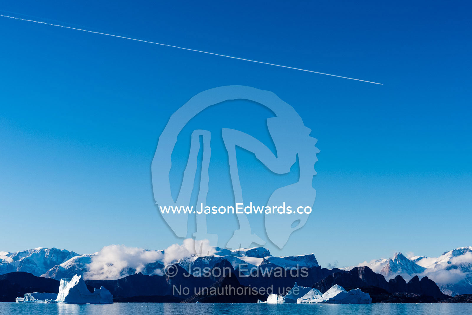 The vapour trails of a passenger jet dissect a clear blue arctic sky above jagged mountains. Milne Land, Hall Bredning Fjord, Scoresby Sound, Greenland