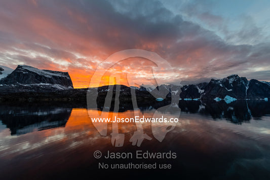 A flaming sunset casts pastel light across storm clouds in a rugged arctic fjord. Milne Land, Mudderbugt, Bjorne Islands, Scoresby Sound, Greenland