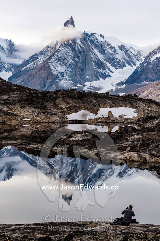 A hiker photographs a jagged spire of rock shrouded in ice and snow reflecting in an arctic pool. O Fjord, Island Fjord, Milne Lan, Grundtvigskirken, Bjorne Islands, Scoresby Sound, Greenland
