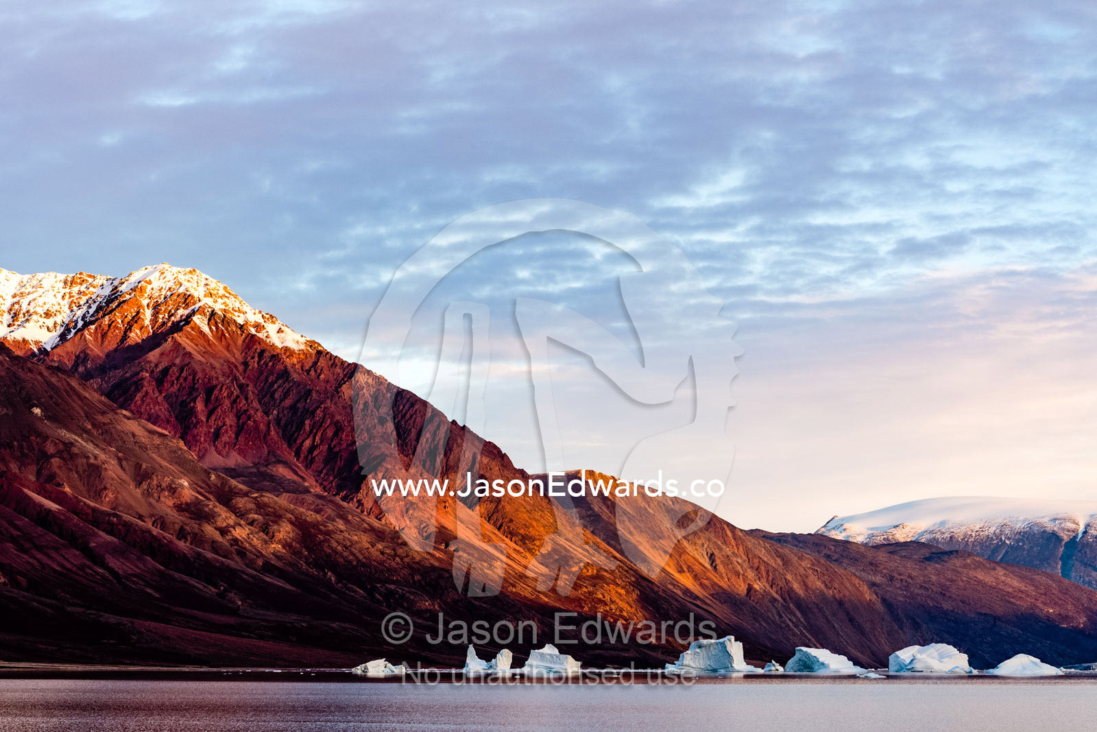 Dawn casts a blood-red glow over barren Arctic mountains surrounded by icebergs. Scoresby Sound, Greenland