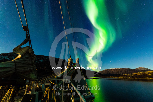 A snake-like ribbon of iridescent green northern lights above the bow of a Tall Ship Schooner. Renodde, Rode Fjord, Scoresby Sound, Greenland