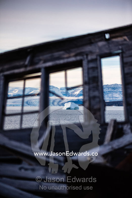 A weathered timber wall and window of a seal hunters hut looks over an iceberg in the arctic. Denmark Island, Hekla Havn, Scoresby Sound, Greenland