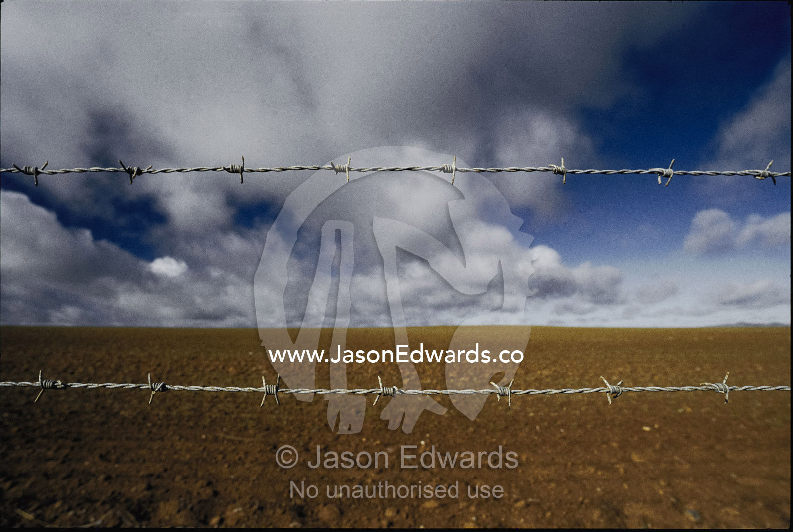 A vast, desolate landscape with dramatic clouds is intercepted by barbed wire fence. Northwestern Victoria, Australia.
