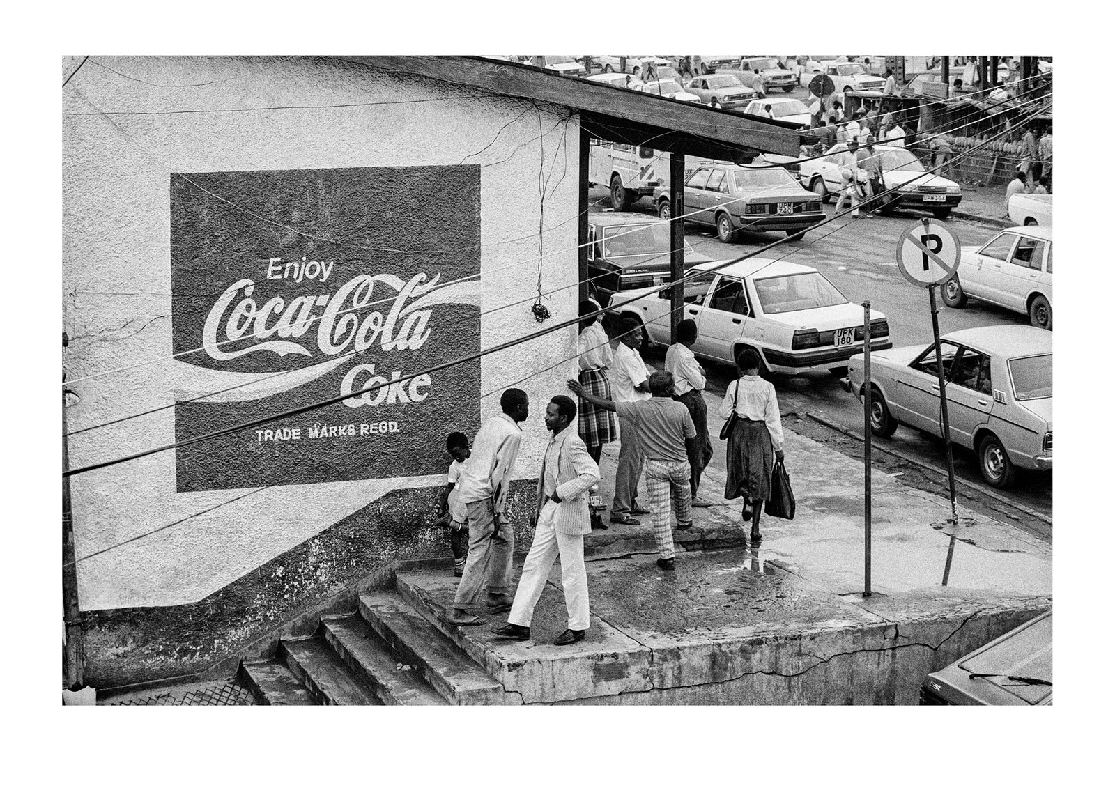 People gather on a busy street corner beneath a Coca Cola sign in downtown Kampala. Captured on Ilford HP5 black and white negative film. Kampala, Western Uganda.